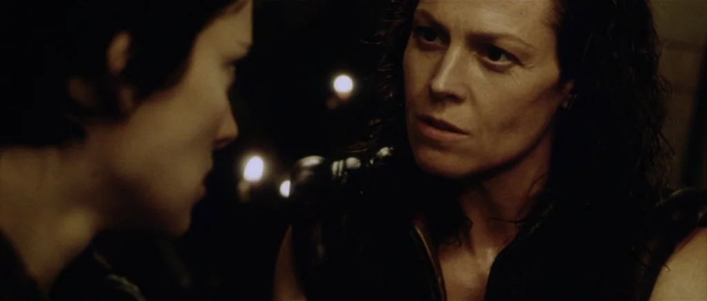 Alien: Resurrection (1997) movie still: medium — A dark, dimly lit environment with out-of-focus light sources in the background. Ellen Ripley looks…