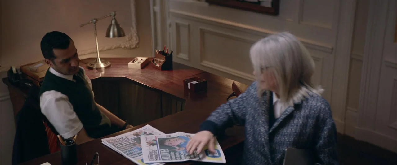 Hampstead (2017) movie still: medium — An interior reception area features a dark wood desk and white panelled walls. A woman hands a newspaper to a…
