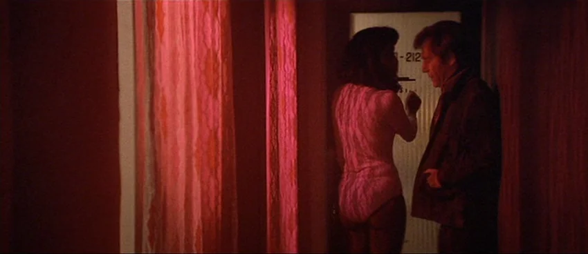 California Split (1974) movie still: medium — A dimly lit motel corridor is saturated in deep red light from outside. A woman in a leotard talks to…