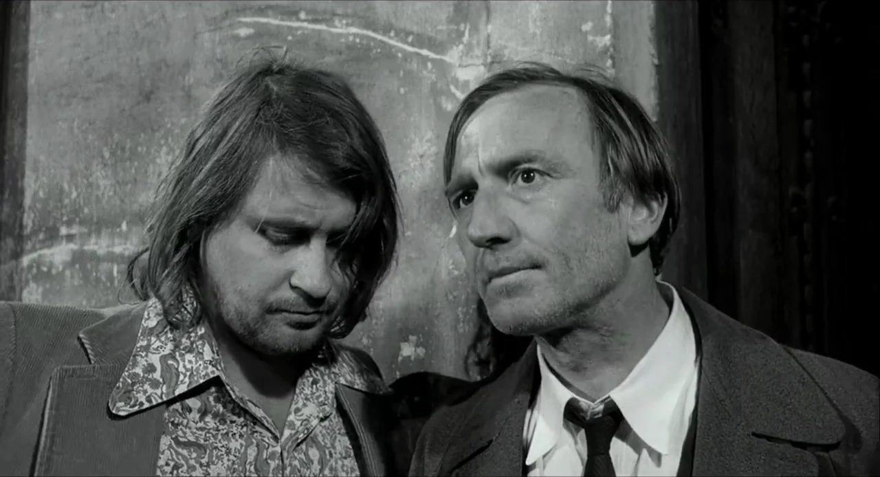 The Bohemian Life (1945) movie still: medium — Two men stand near a textured interior wall surface in a plain hallway. The younger man looks…