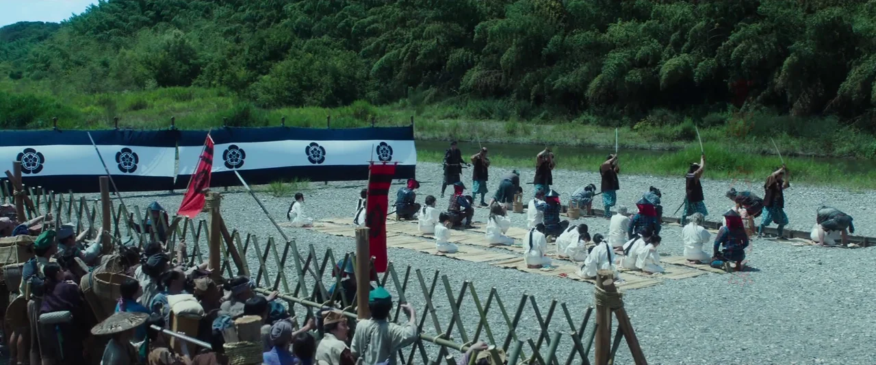 Kubi (2023) movie still: wide — A rocky riverbank features a crowd gathered behind a wooden crisscross fence. In the center, people in historical…