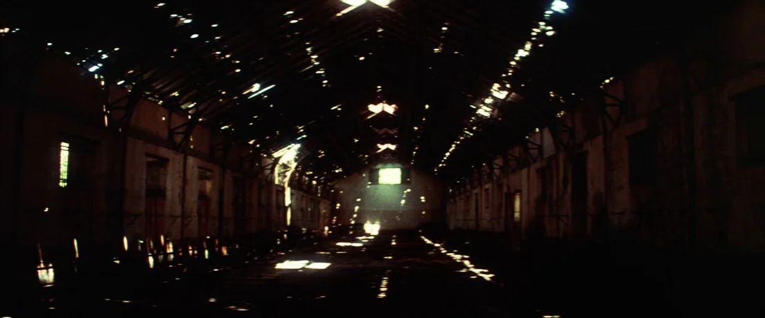 Papillon (1973) movie still: establishing — A dark, empty industrial building has structural beams and holes in the roof. Multiple shafts of…