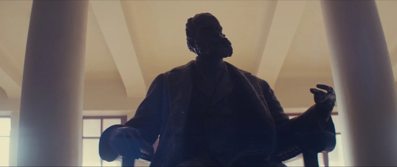Neruda (2016) movie still: medium — A bronze statue of a man sits between two large pillars in a bright room. The man faces forward with one hand…