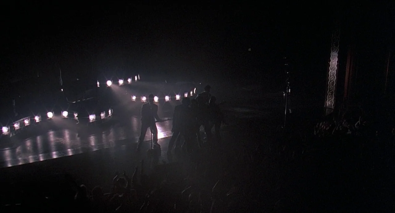 Streets of Fire (1984) movie still: wide — A dark concert stage with a band performing behind a curved line of lights. The audience is visible as a…