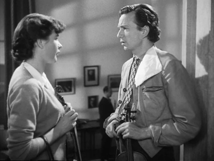 To Joy (1950) movie still: medium — An interior space contains a man and woman holding violins while talking. A third person is faintly visible in…