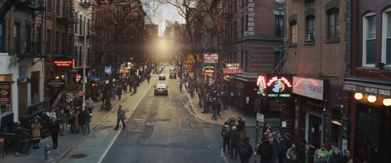 The Sun Is Also a Star (2019) movie still: wide — A bustling city street lined with brick buildings and multiple neon storefront signs. Pedestrians…