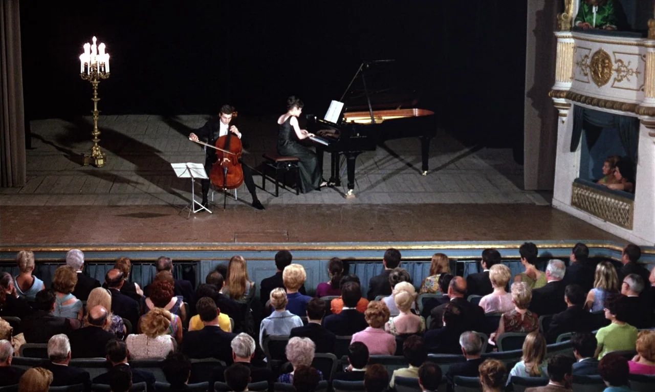 The Bride Wore Black (1968) movie still: wide — A wooden performance floor holds a man on a cello and a grand piano. An audience sits in rows of…