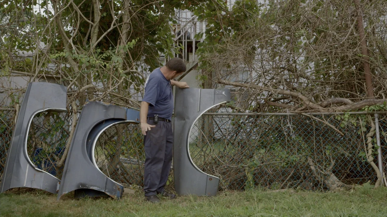 The Last Race (2022) movie still: medium — A man stands in a grassy backyard next to a chain-link fence overgrown with vines. The subject examines…