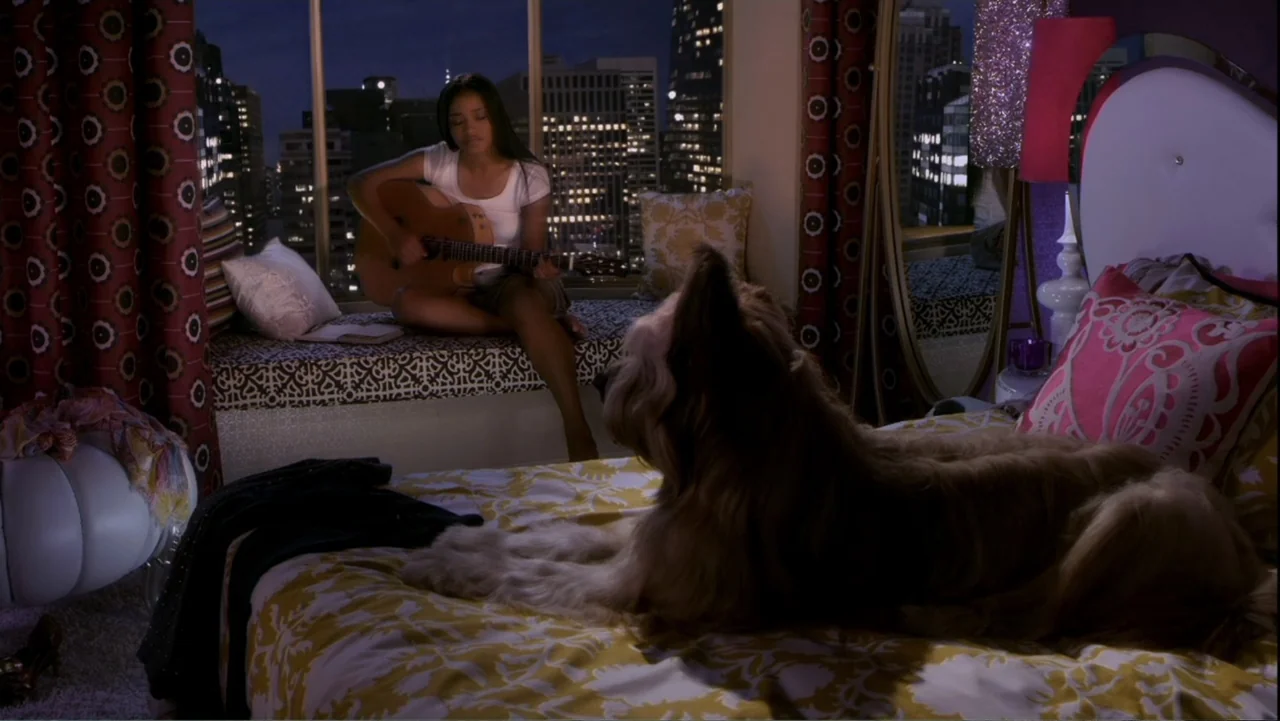 Rags (2012) movie still: medium — A modern bedroom with a large window overlooking a illuminated city at night. A young woman sits on a window bench…