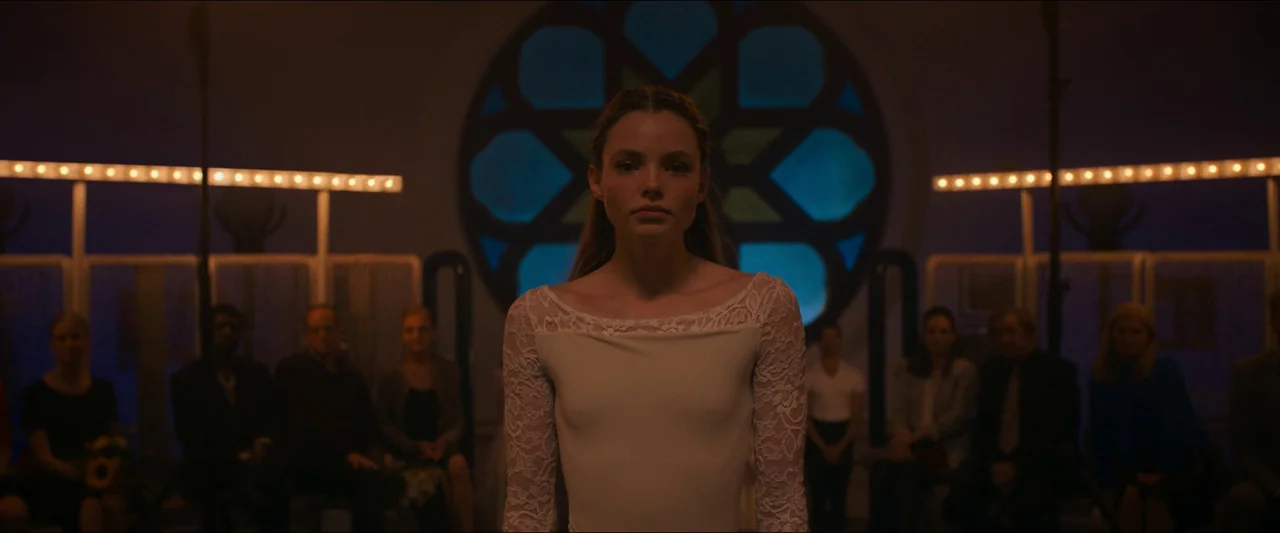 Birds of Paradise (2021) movie still: medium — A dimly lit room features a large circular stained glass window in the center. A young woman in a…