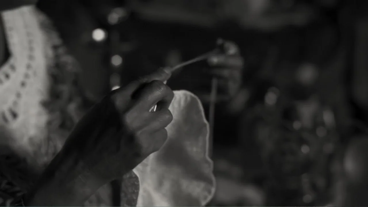 Faya Dayi (2021) movie still: closeup — A close view of hands working with delicate lace in a dimly lit space. Fingers hold a needle and thread…