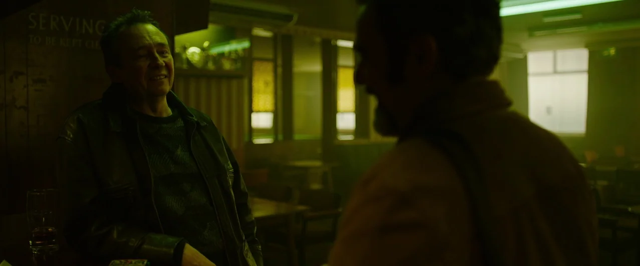 Ghost Stories (2017) movie still: medium — A dimly lit, green-tinted pub interior with blurred tables and chairs in the background. Two middle-aged…