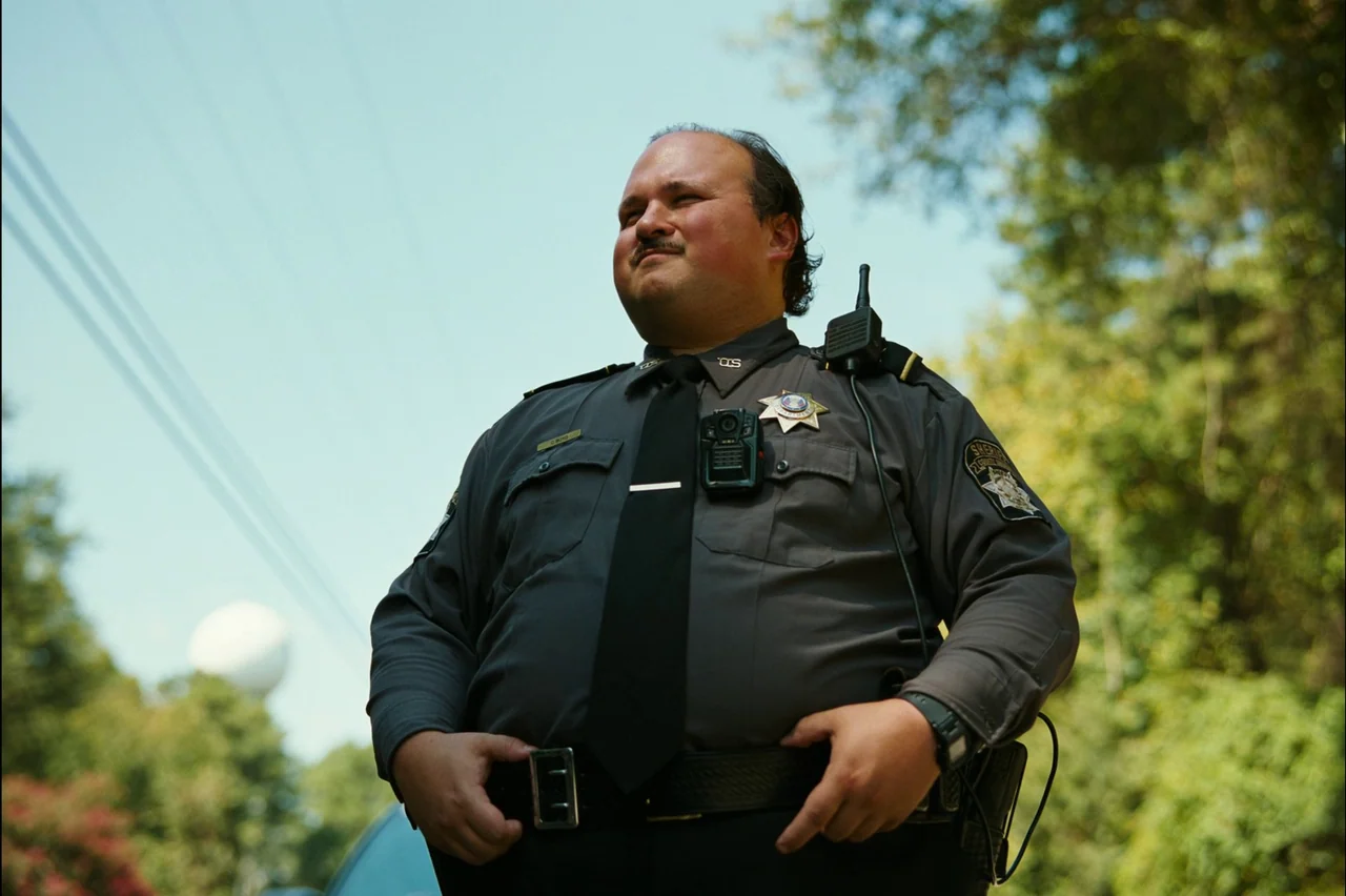 Bugonia (2025) movie still: medium — An outdoor roadside setting with a bright sky and green trees in the background. A police officer stands in his…