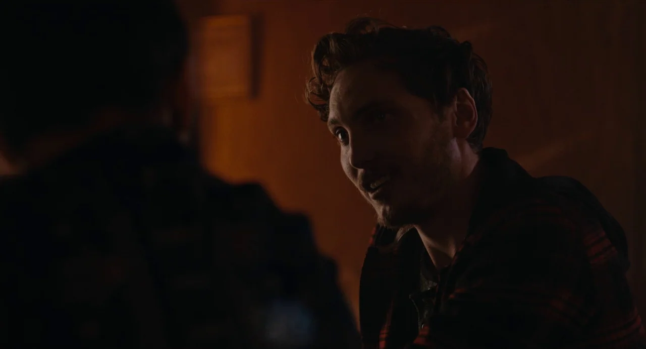 Lingua Franca (2019) movie still: closeup — A dark indoor space with wooden textures and indistinct background elements. A man in a plaid hoodie…