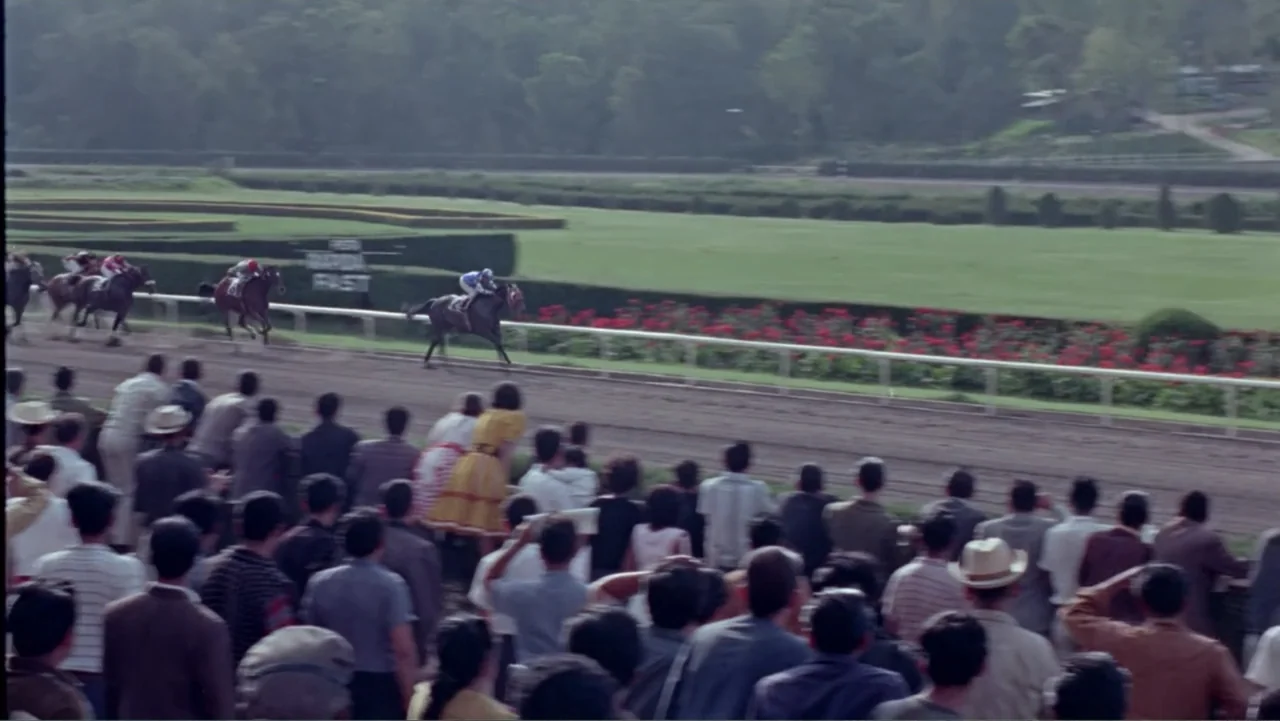 Los Dos Rivales (1966) movie still: wide — A crowded outdoor racetrack features a long dirt track with green fields beyond. A group of spectators in…