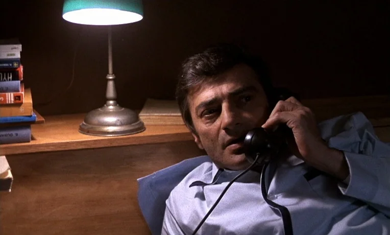 The Man Who Loved Women (1977) movie still: medium — A darkened room features a wooden shelf with a small lamp and books. A man wearing a light blue…