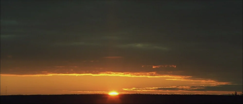 The Straight Story (1999) movie still: establishing — A vast dark landscape meets a heavy cloud cover at the horizon line. The sun emits a bright…