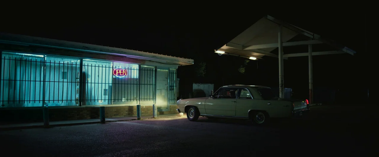 LaRoy, Texas (2023) movie still: wide — A quiet gas station at night is lit by a vibrant red neon sign. A vintage yellow car is parked in the dark…