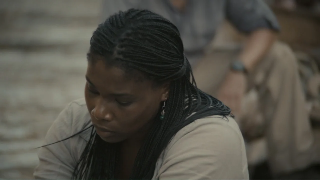 The Dinosaur Project (2012) movie still: medium — A woman with braided hair sits outdoors during the day in a neutral setting. She looks down while…