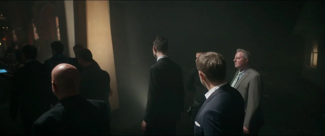 Utopia (2013) movie still: medium — A dark, hazy interior space is occupied by several men in formal business suits. The group faces away from the…