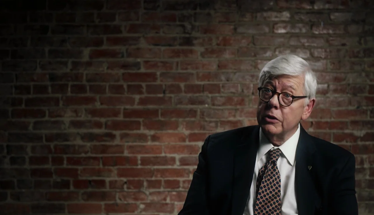 13th (2016) movie still: medium — An elderly man is positioned against an exposed red brick wall. He sits in a suit while engaged in an interview.