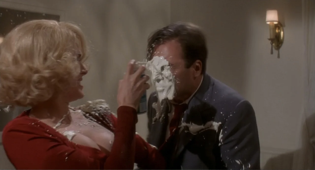 Fatal Instinct (1993) movie still: medium — A well-lit indoor space featuring a wall-mounted brass sconce and plain off-white walls. A woman in a…