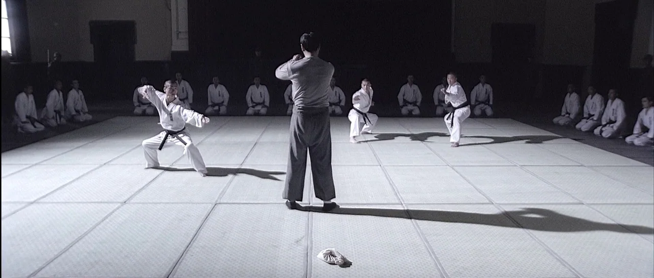 Ip Man (2008) movie still: wide — A spacious room with a matted floor and students kneeling against the dark walls. An instructor stands in the…