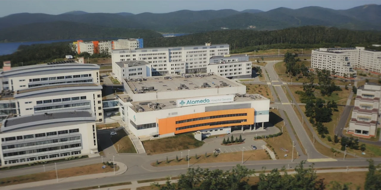 Our Fault (2025) movie still: establishing — A modern hospital complex sits nestled in a landscape of green hills and trees. Several multi-story…