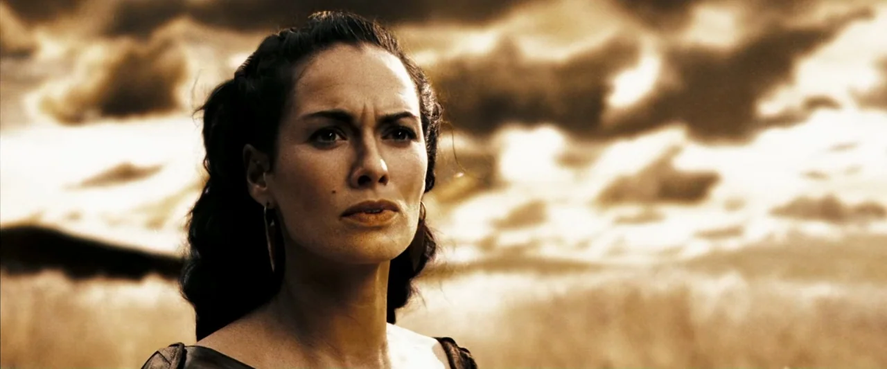 300 (2006) movie still: closeup — A woman stands in a golden field under a sky filled with heavy clouds. She looks off-camera as the bright sun…