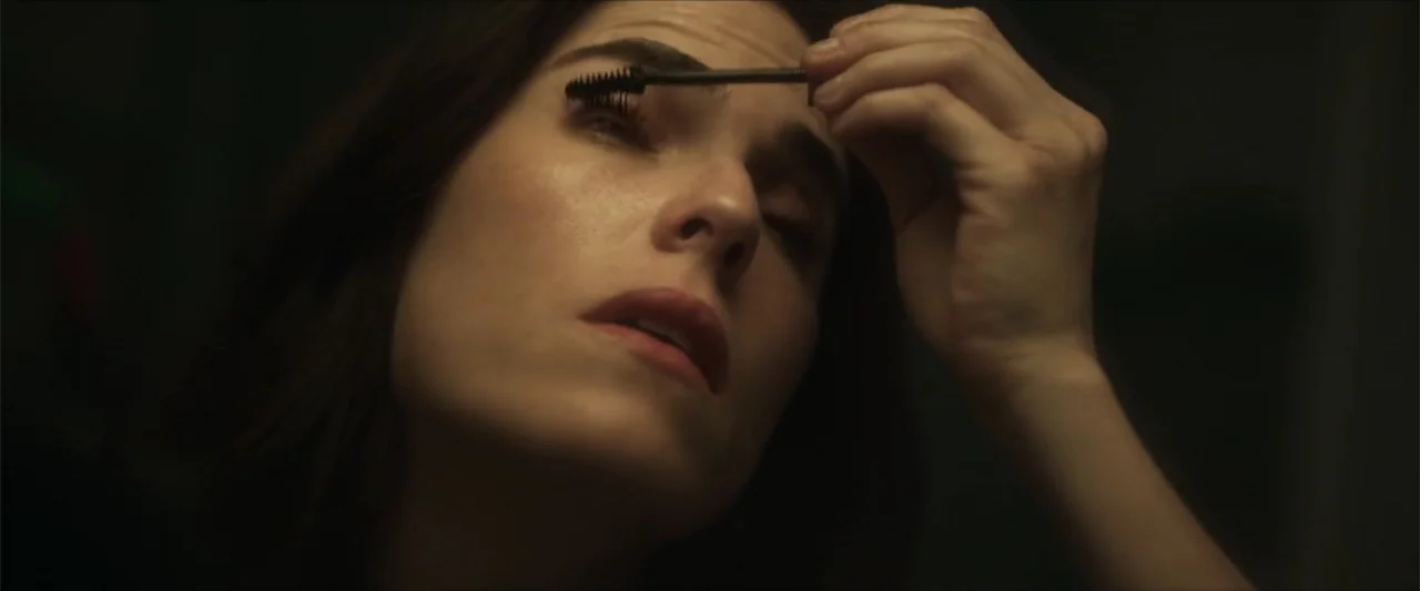 Dive (2013) movie still: closeup — A dimly lit room with muted neutral walls. A woman holds a mascara wand and carefully applies makeup to her lashes.