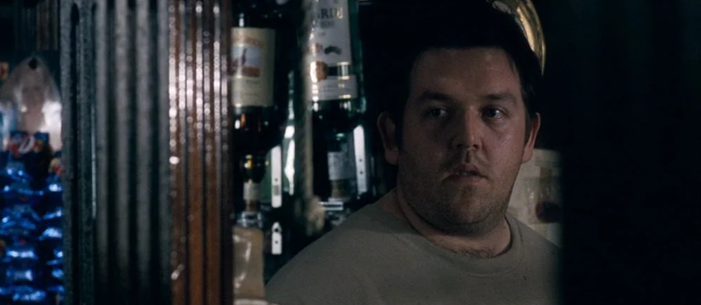 Shaun Of The Dead (2004) movie still: medium — A dimly lit pub interior features dark wooden shelving lined with various liquor bottles. A man in a…