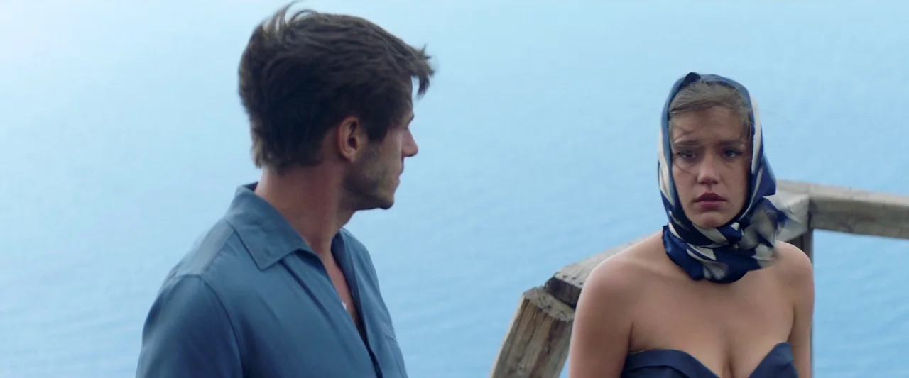 Sibyl (2019) movie still: medium — A wooden railing overlooks a vast, calm body of bright blue water. A young man looks at a woman wearing a blue…