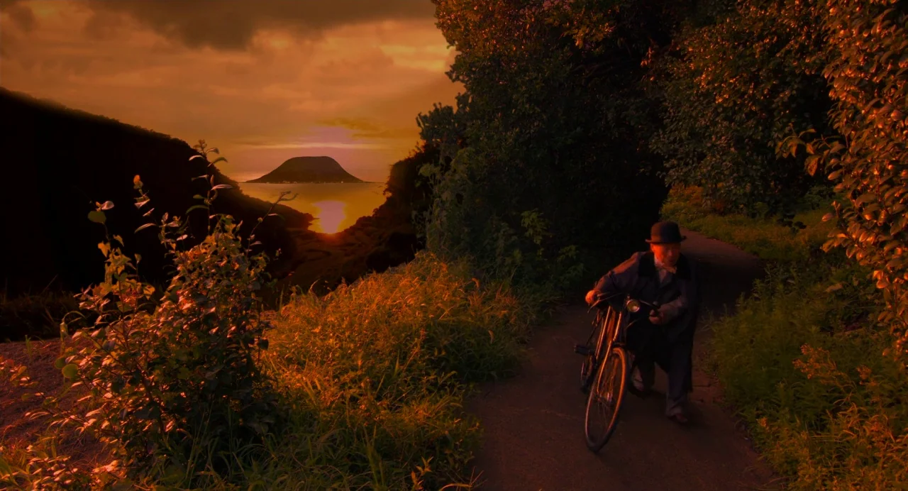 Hanagatami movie still: wide — A rural coastal path winds through thick vegetation during a bright sunset. An older man in a hat walks while pushing…