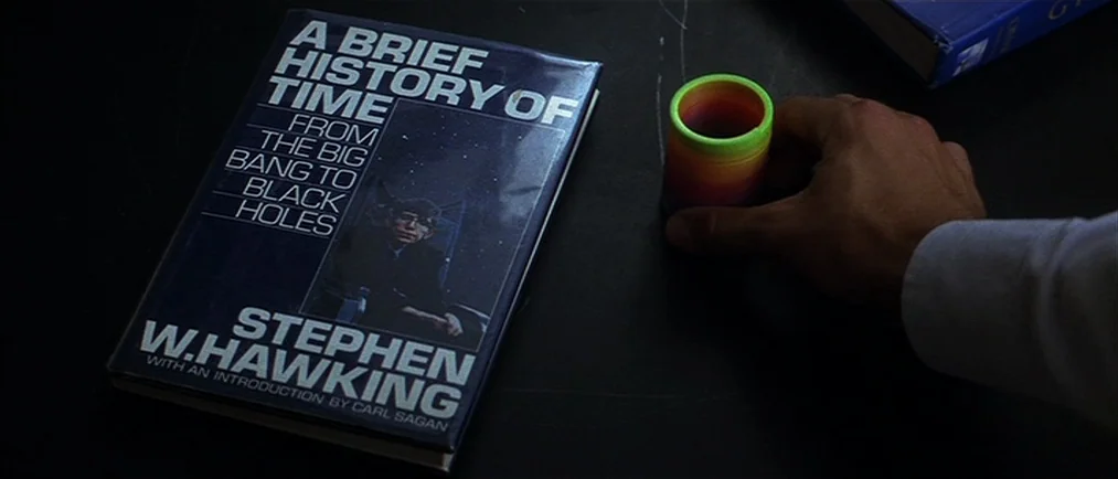 Donnie Darko movie still: closeup — A dark surface holds a copy of A Brief History of Time by Stephen Hawking. A hand holds a colorful plastic…