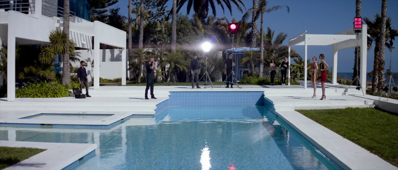 The Neon Demon (2016) movie still: wide — A modern white house terrace features a large blue swimming pool surrounded by palm trees. A film crew…