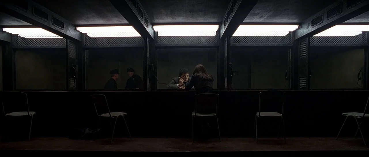 Dark City (1998) movie still: wide — A dimly lit visiting area in a prison with individual glass partition booths. A man and a woman sit in separate…