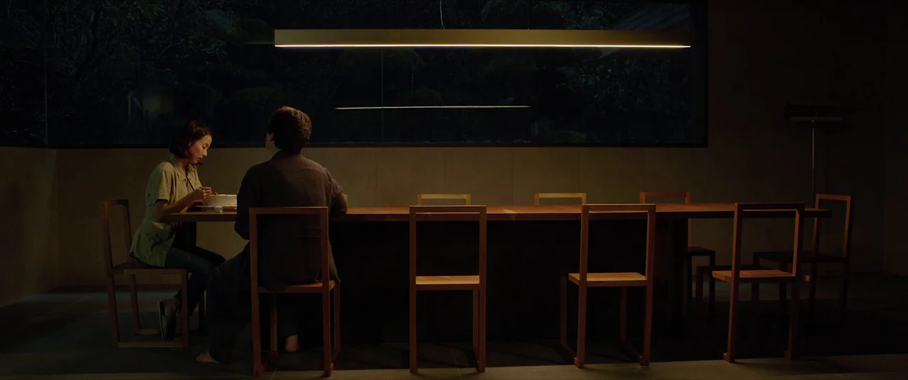 Parasite (2019) movie still: wide — A minimalist dining room features a long wooden table with several matching wooden chairs. A man and a woman sit…