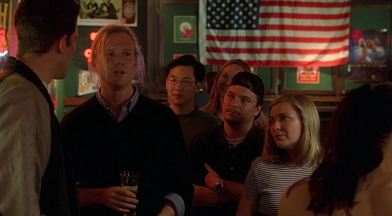Good Will Hunting (1997) movie still: medium — A dark bar interior features an American flag hanging on a green wall. A group of young adults stands…