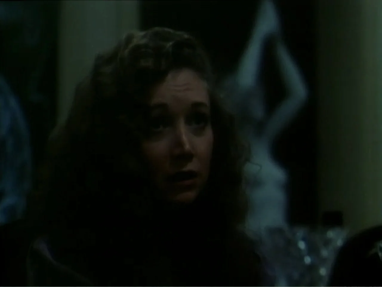Curtains (1995) movie still: closeup — A dimly lit indoor space featuring dark furniture and a blurry white figurine. A woman with wavy hair gazes…