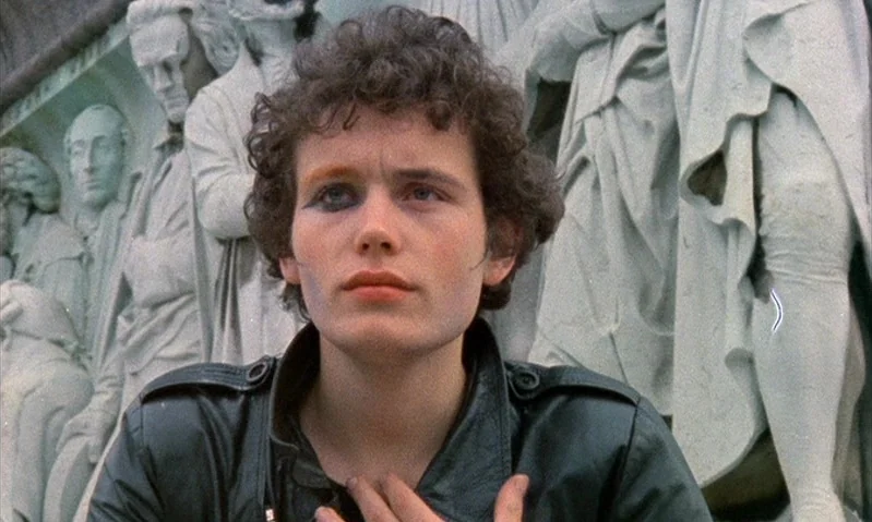 Jubilee (1978) movie still: medium — A person stands before a large, carved stone relief of multiple human figures. The individual looks upward with…