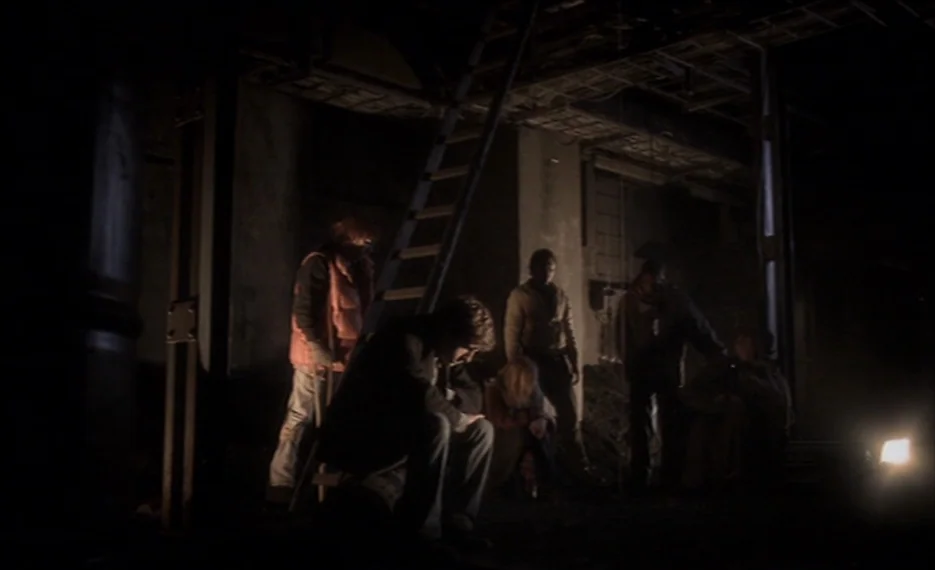 Dear Wendy (2005) movie still: wide — A dark, unfinished basement with exposed ceiling rafters and rough concrete walls. Several people gather in…