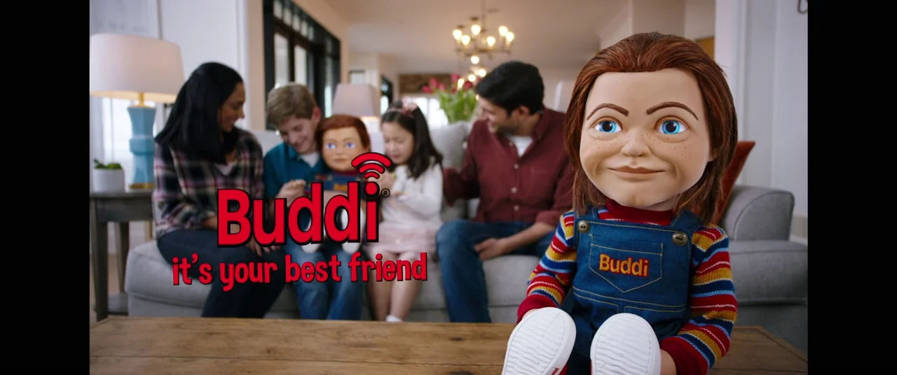 Child's Play (2019) movie still: medium — A well-lit contemporary interior with a family sitting on a couch in the background. A Buddi doll sits in…