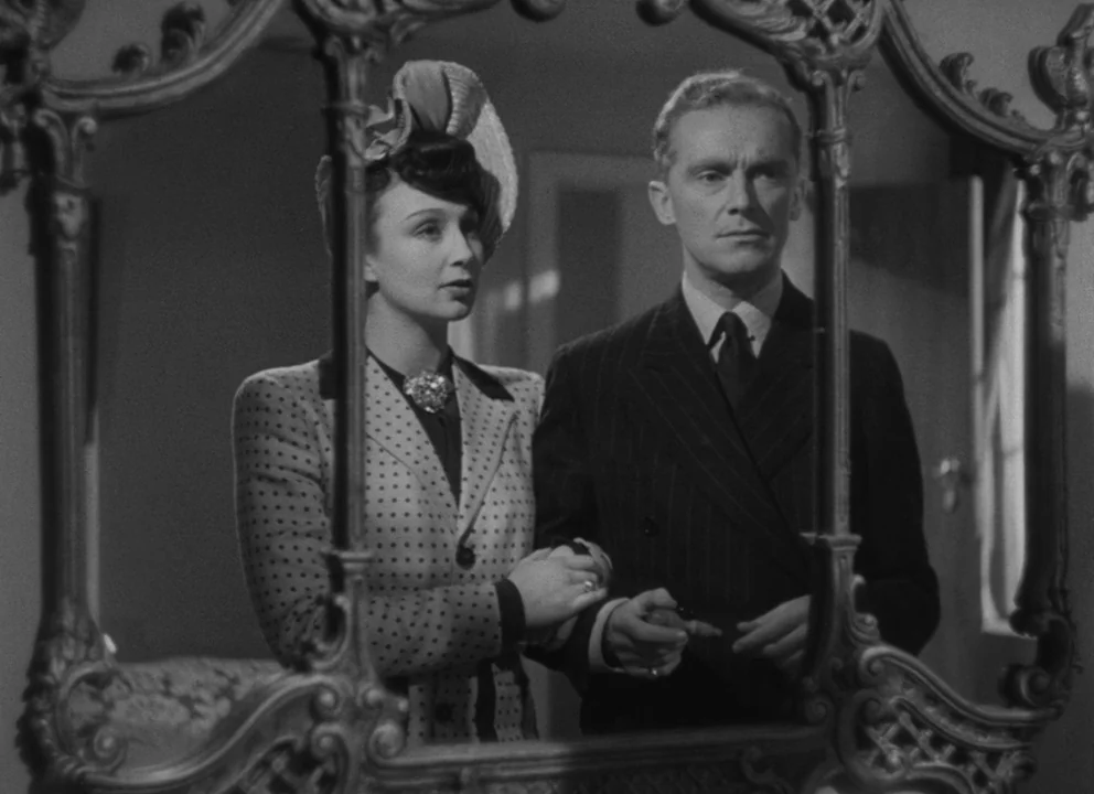 Dead of Night (1945) movie still: medium — A man and a woman in formal attire stand together inside a room. They gaze directly into an ornate,…