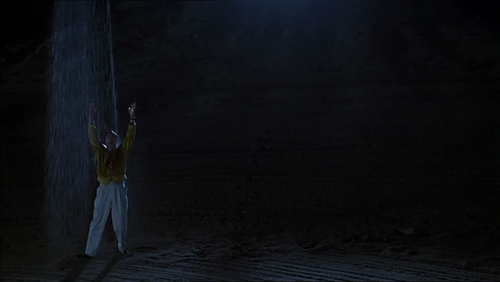 The Truman Show movie still: wide — A dark, cavernous space features textured stone walls and a sandy ground. A man stands beneath a narrow stream…