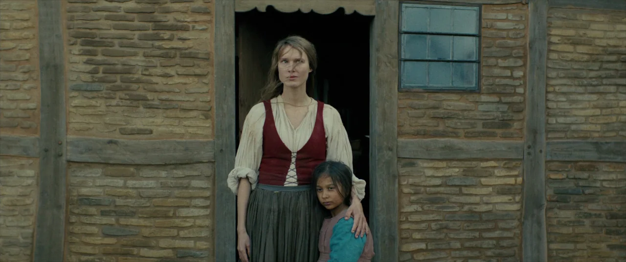 The Promised Land (2023) movie still: medium — A rustic building constructed of rough bricks and wooden timber frames stands behind them. A woman…