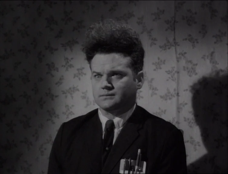Eraserhead (1977) movie still: medium — A dimly lit interior with light floral wallpaper providing the background. A man stands facing forward…