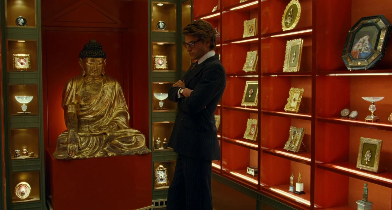 Saint Laurent (2014) movie still: medium — A room with red lacquered walls and shelves filled with ornate framed objects and antiques. A man in a…