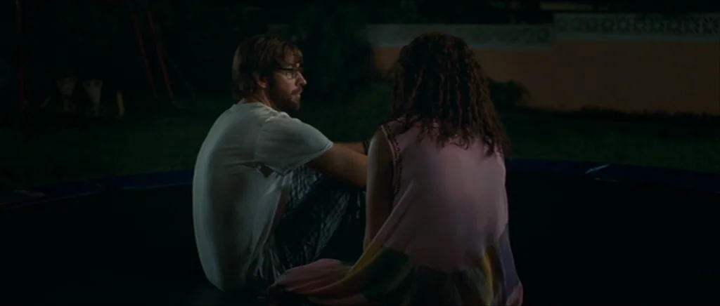 Away We Go (2009) movie still: medium — A man and woman sit together on a dark backyard trampoline at night. The surrounding yard is dim and…
