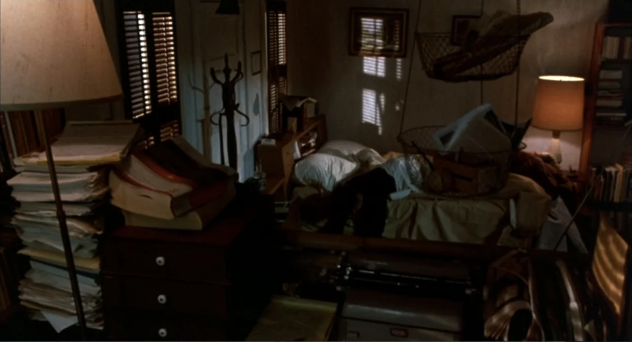 Murphy's Law (2009) movie still: wide — A cluttered room contains stacks of papers, a typewriter, and a bed near a window. A person is lying prone…