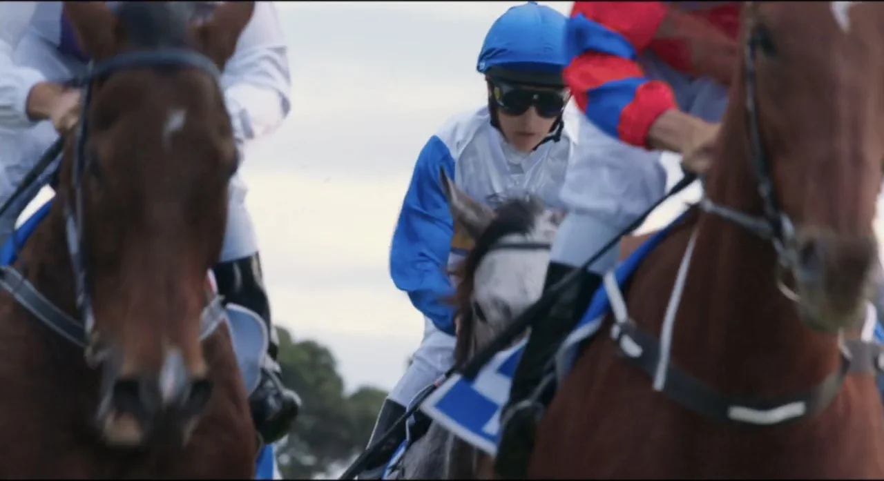 Ride Like a Girl (2019) movie still: medium — A group of jockeys rides horses at full gallop on a dirt track. The horses are in tight proximity…